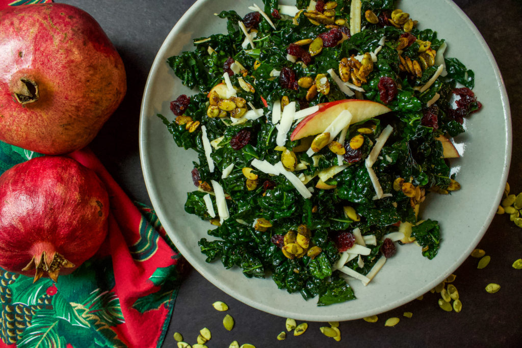 kale salad with cheese, pumpkin seeds, cranberries, and apple next to pomegranates on the table